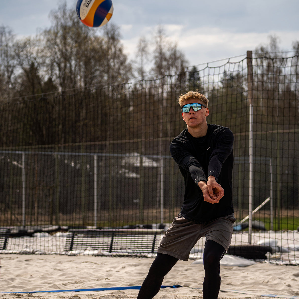 Samu Pulkkinen kaivamassa palloa beach volley -kentällä, hiekkaa ja lehdettömät puut taustalla